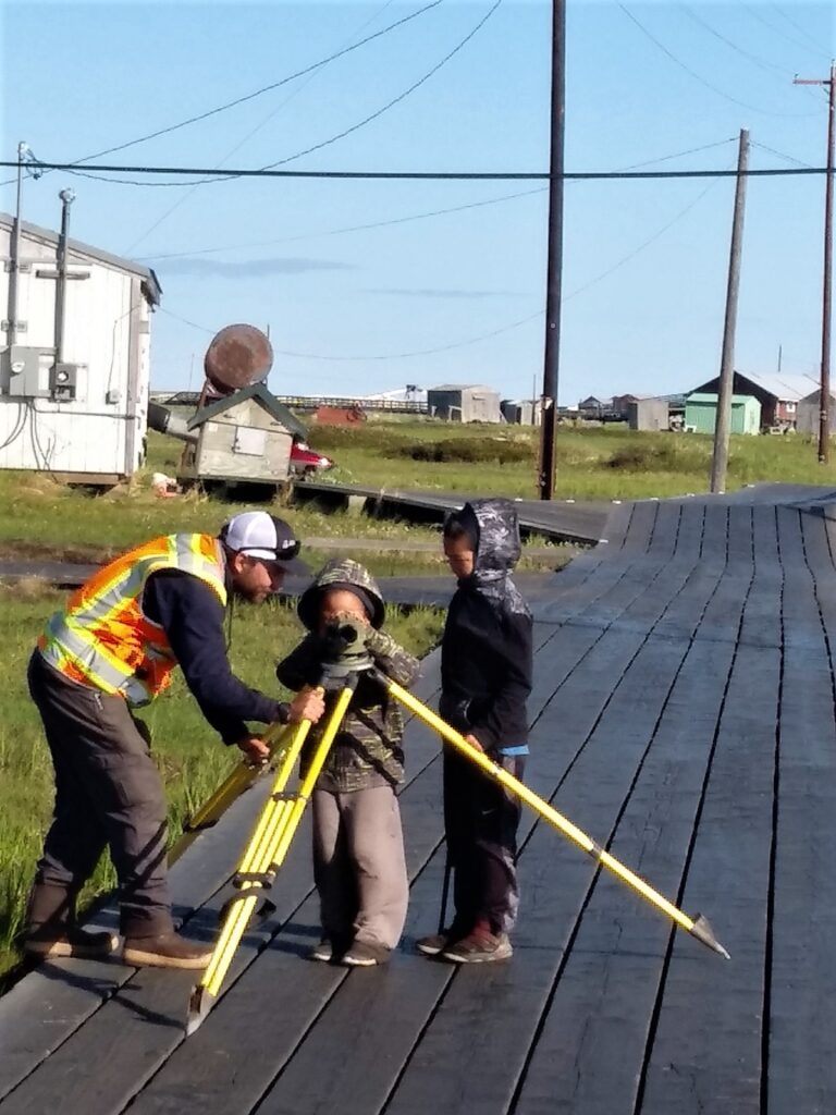 Land surveyor demonstrating a tripod-mounted surveying instrument to two children on a boardwalk in a rural Alaska community.