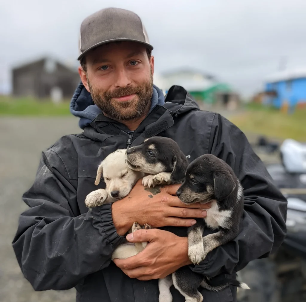 Drew holding three young puppies in his arms, smiling outdoors while wearing a dark jacket and cap, with a rural Alaskan setting in the background.
