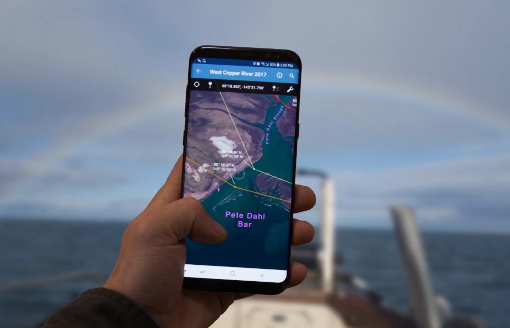 cHand holding a smartphone displaying a georeferenced nautical map on a mapping app, with open ocean and the bow of a boat visible in the background.
